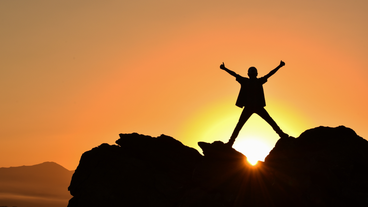 Mensch auf einem Berg streckt die Arme in die Luft, dahinter ein Sonnenaufgang